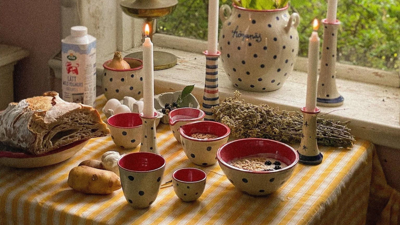 Handmade ceramic polka dot tableware set with red interior, artisan bread, and lit candles on yellow striped tablecloth
