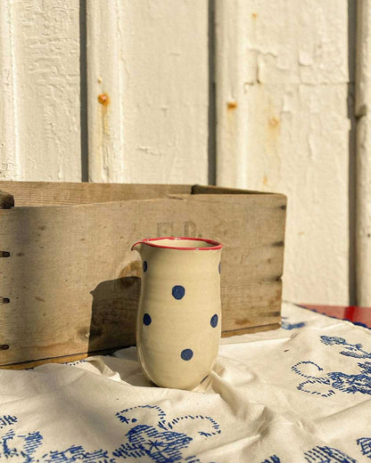 Handcrafted Spanish ceramic large milk jug with blue polka dots and red rim, displayed on vintage blue-patterned textile beside wooden crate