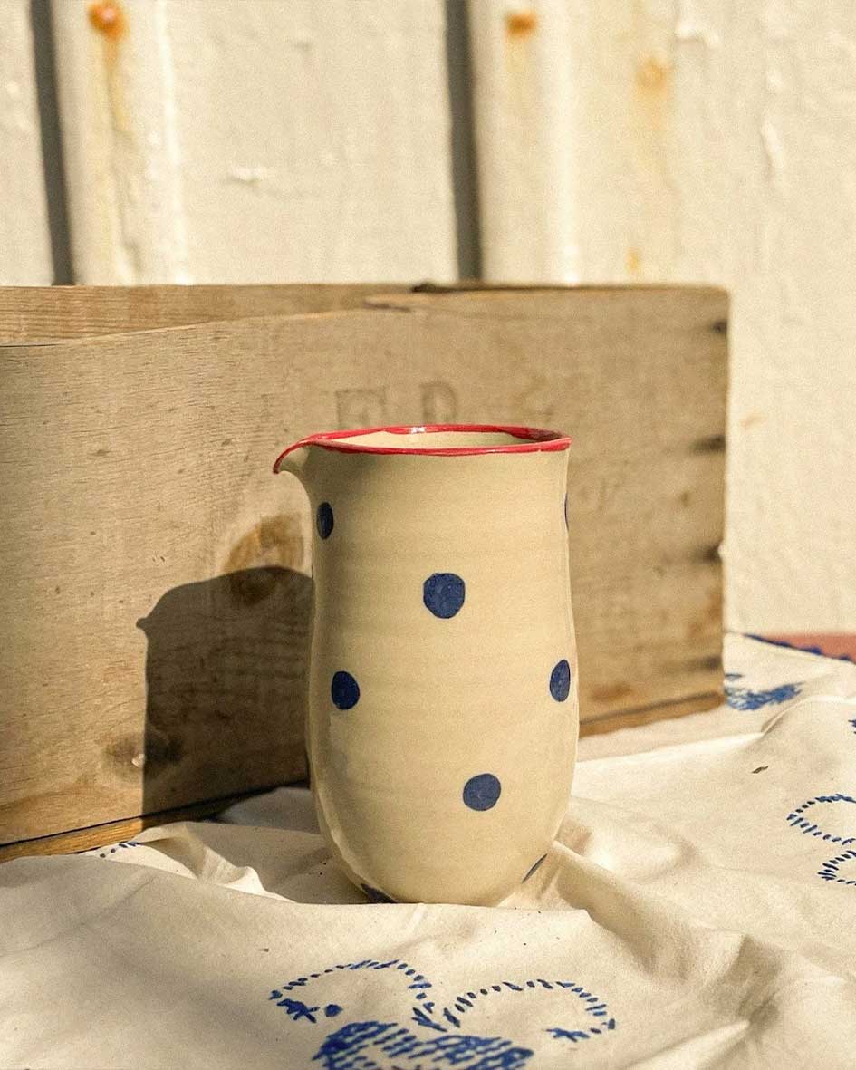 Handcrafted Spanish ceramic small milk jug with blue polka dots and red rim, resting on vintage blue-patterned textile