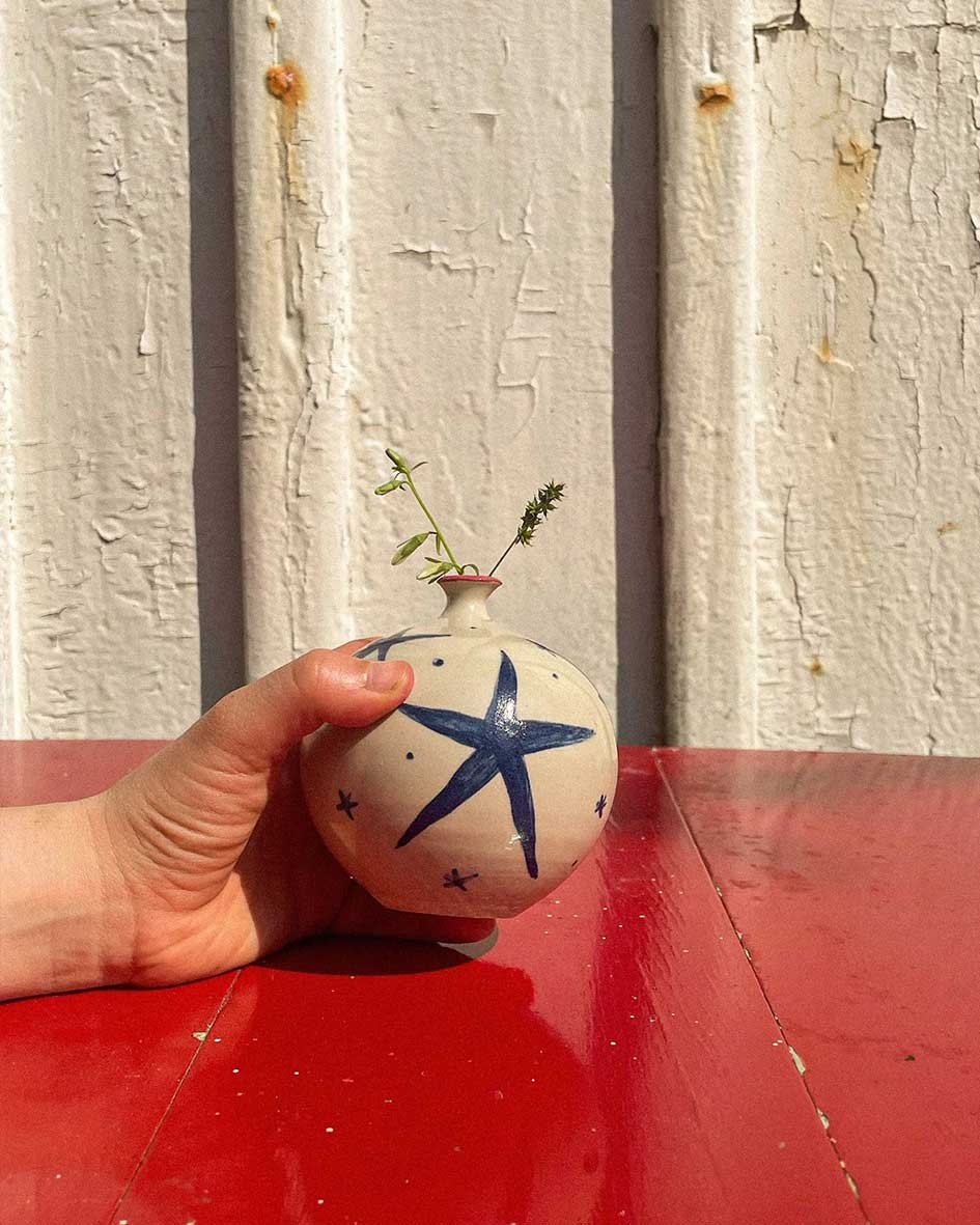 Handcrafted Spanish ceramic small vase with blue star design, held against rustic white wall with delicate botanical accent